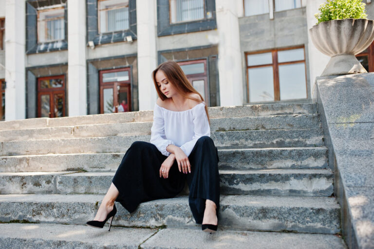 Une femme assise sur un escalier devant une grande maison, en regardant les talons qu'elle porte avec son pantalon velours