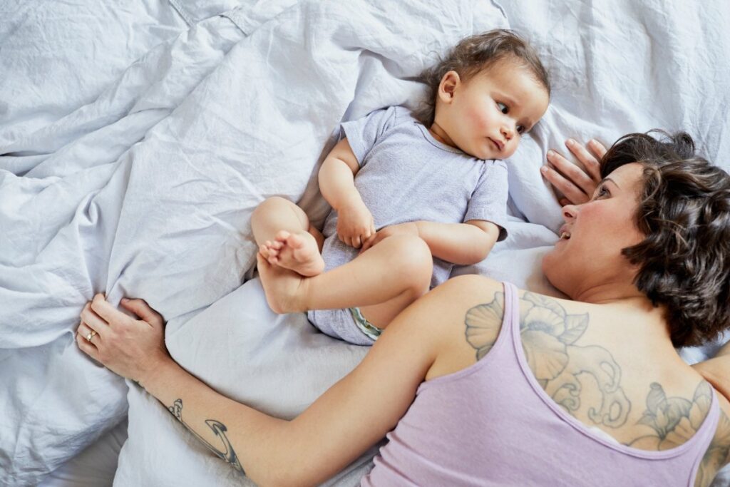 Une maman allongée sur le lit avec plein de tatouage sur son dos et qui joue avec son bébé