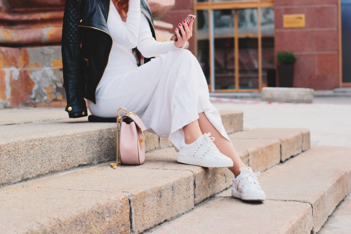Une femme assise sur un escalier avec une robe longue blanche, une veste noire et un basket blanc