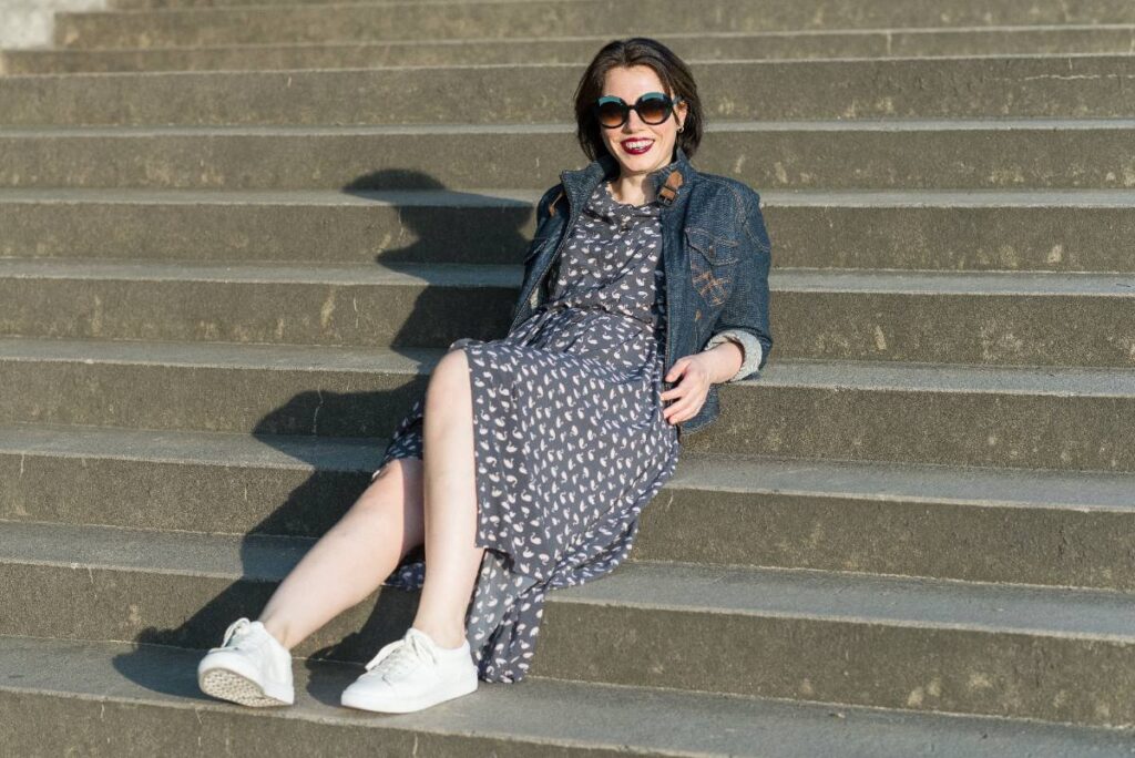 Une femme assise sur un escalier en portant une robe longue, une lunette noire et un basket blanc